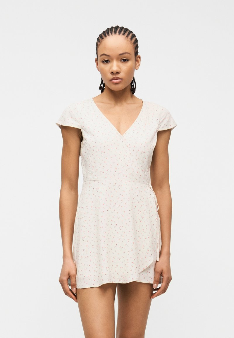 Γυναίκα με πλεξούδες, φορώντας ένα κοντό λευκό φόρεμα τύπου wrap με μικρό κόκκινο λουλουδάτο σχέδιο, στέκεται μπροστά σε απλό φόντο.