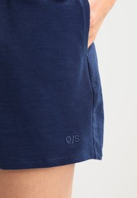 Ruka v kapse na tmavě modrých texturovaných šortkách s malým logem "Q/S" blízko lemu na jednobarevném světlém pozadí.