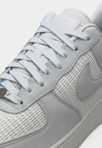 Pantof sport alb și gri deschis cu partea superioară din plasă, suprapuneri din piele întoarsă, șireturi asortate și o emblemă vizibilă a mărcii pe lateral.