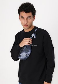Blauwe tie-dye schoudertas met een gestructureerde afwerking, verstelbare schouderband en ritssluiting, gedragen over een zwarte sweatshirt.
