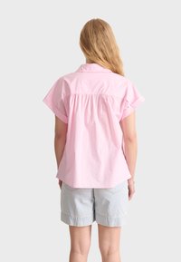Una camicia di cotone rosa con vestibilità rilassata, maniche corte, yoke posteriore con arricciature e orlo curvo. Indossata con pantaloni shorts in denim azzurro chiaro.