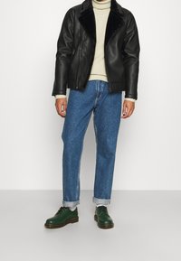 Czarna skórzana kurtka z futrzanym kołnierzem, beżowy sweter z golfem, niebieskie dżinsy, zielone skórzane buty z ciemną podeszwą.