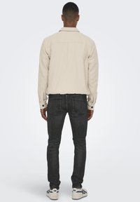 Jachetă bej cu buton la mansetă, asortată cu blugi negri. Outfitul are un design curat și minimalist.