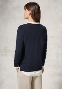 Marineblauwe gebreide trui met lange mouwen, een ronde hals en geribbelde accenten, gelaagd over een wit overhemd met kraag. Zachte textuur.