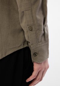 Gros plan d'une manche de chemise en velours côtelé beige avec un poignet boutonné sur le bras d'une personne, sur un fond clair uni.