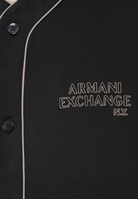 Μαύρο πουκάμισο με κουμπιά, λευκή περιθωριοποίηση και κεντημένο κείμενο "ARMANI EXCHANGE N.Y." στην αριστερή πλευρά του στήθους.