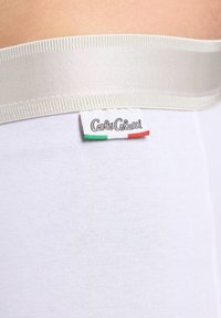 Close-up van een witte elastische tailleband op witte stof met het label "Carlos Colucci" met groen, wit en rode strepen.