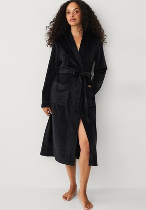 Vrouw met krullend haar, gekleed in een zwarte fluwelen badjas die rondom de taille is gestrikt, met één hand in de zak, staand op blote voeten tegen een eenvoudige achtergrond.