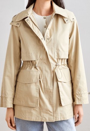 Femme portant une veste utilitaire beige avec poches avant, taille à cordon, col rabattu et poignets boutonnés sur un pull côtelé blanc.