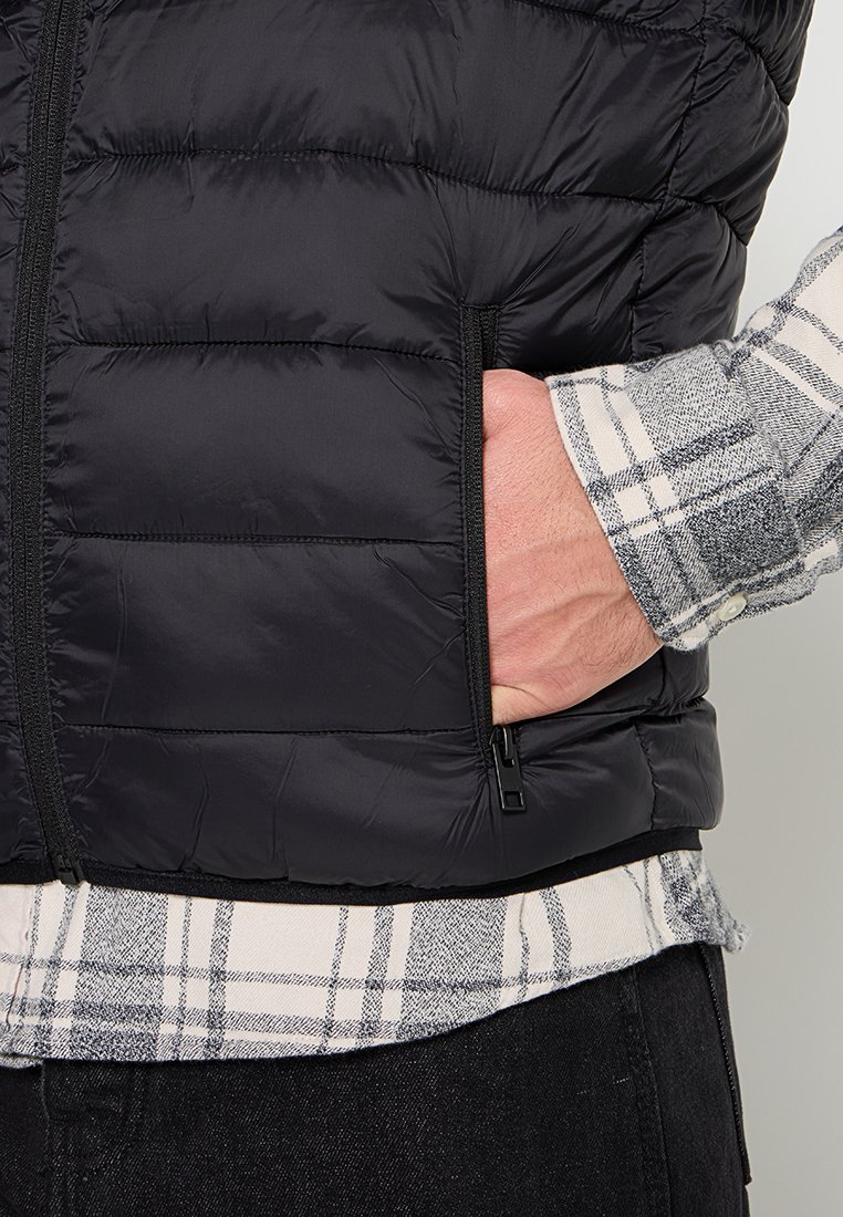 Svart puffväst med horisontellt quiltat mönster, dragkedjefickor och en slät textur. Under finns en grå och vit rutig skjorta synlig.