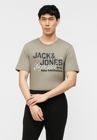 Homme portant un T-shirt vert olive avec le texte "JACK & JONES Superior New Aesthetics", associé à un pantalon noir, debout devant un fond blanc.