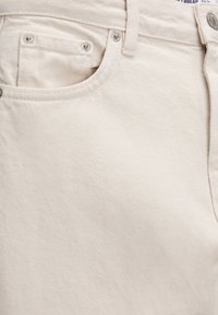 Zbliżenie na kremowe dżinsy z denimu pokazujące przednią kieszeń, kieszonkę na drobne z nitami, pętlę na pasek oraz detale zamknięcia na guzik.