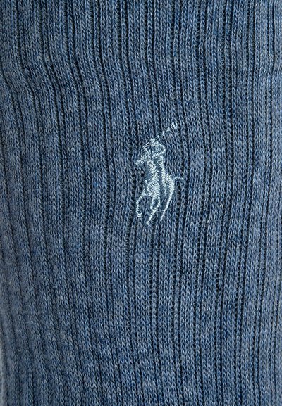 Zilā ribota adīta zeķe ar izšūtu logotipu, kurā attēlots zirgskārs unicorns. Tekstūra šķiet maiga, un krāsa ir vienmērīga visā virsmā.
