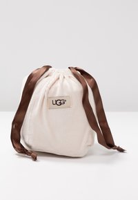 Plátěný sáček s tkaničkou v krémové barvě s žebrovanou strukturou, zdobený hnědými saténovými stuhami a štítkem UGG na přední straně.