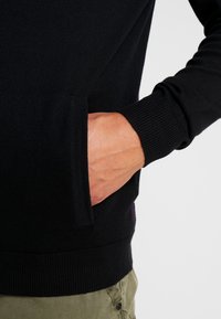 Black sweatshirt with ribbed cuffs and a side pocket. Smooth texture and fitted design. Visible stitching detail near the hem.