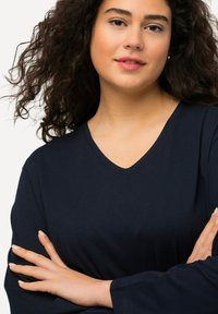 Navy V-Ausschnitt-Oberteil mit langen Ärmeln, aus weichem Stoff gefertigt. Bietet eine lockere Passform und subtile Nähdetails. Modell posiert mit verschränkten Armen.
