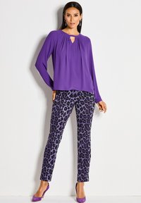 Vrouw in een paarse blouse met lange mouwen, paarse en zwarte luipaardprintbroek en paarse hoge hakken, staand tegen een witte muur.