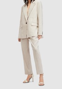 Beżowy lniany garnitur z oversize'ową marynarką i prostymi spodniami. Zauważalne detale to głęboki dekolt oraz jasne sandały na wysokim obcasie.