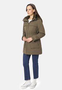 Parka verde oliva con capucha forrada de piel, cierre de cremallera, y dos grandes bolsillos en la parte delantera, llevada sobre jeans de mezclilla azul y zapatillas blancas de caña alta.