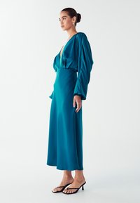 Žena ve smaragdově modrých saténových dlouhých šatech s hlubokým výstřihem do V, stojící v profilu, obutá v černých sandálech na vysokém podpatku a nosící zlaté kruhové náušnice.