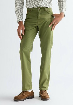 Uomo che indossa pantaloni verde oliva a gamba dritta, scarpe di camoscio marroni e una camicia a quadri bianca a maniche lunghe, in piedi contro uno sfondo semplice.
