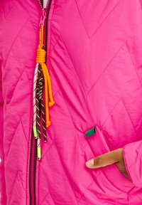 Jasně růžová prošívaná bunda s zipem, barevnými stahovacími šňůrkami s třásněmi a boční kapsou s jemným zeleným akcentem.
