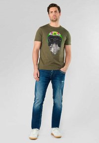 Camiseta de algodón verde olivo con un gráfico de un casco con gafas de sol. Lleva puesta con vaqueros azules desgastados y zapatillas blancas.