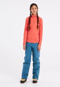 Koraalkleurige top met lange mouwen en een rits aan de voorkant, gecombineerd met losvallende blauwe broek en tan-kleurige laarzen. Vloeiende textuur door het hele outfit.