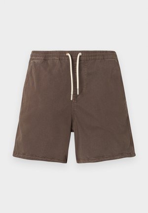 Homme portant un t-shirt vert sauge à manches courtes, un short marron, des chaussettes blanches et des baskets crème, debout devant un fond blanc uni.