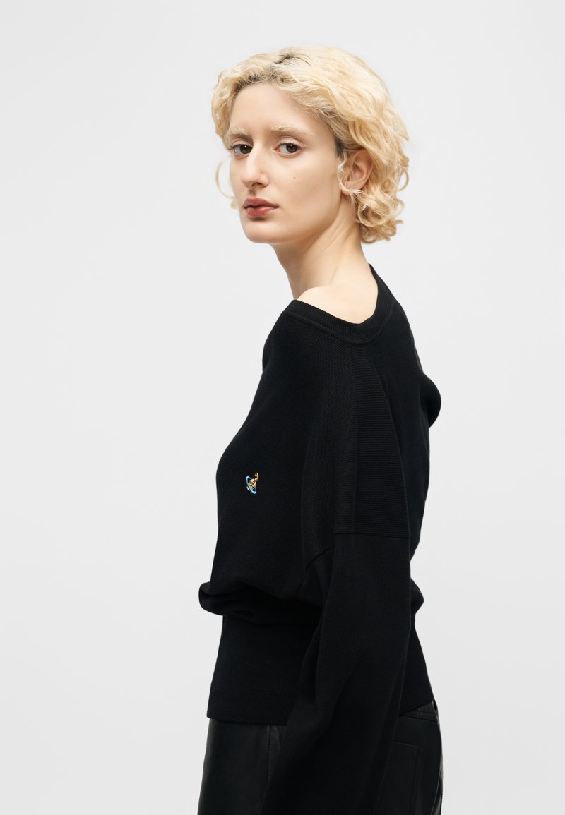 Young person with short curly blonde hair wearing a black sweater with a small colorful planet emblem on the chest, posing sideways.