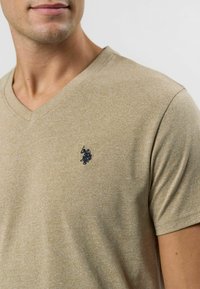 Tricou bej cu decolteu în V, cu o textură moale, având un mic logo bleumarin pe partea stângă a pieptului și mâneci scurte.