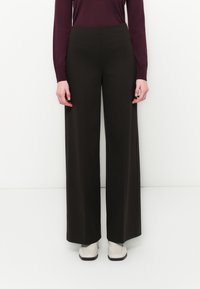 Schwarze weit geschnittene Hosen mit glatter Textur, hoher Taille und minimalistischer Gestaltung. Getragen mit einem dunklen Oberteil und hellen Schuhen.