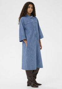 Denim srajčna obleka, svetlo modra, dolgi rokavi, zapiranje na gumb, dva sprednja žepa, dolžina do sredine meč, kombinirana z rjavimi lakastimi škornji do kolen.