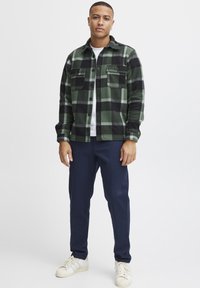 Camisa de franela a cuadros verdes y negros con dos bolsillos en el pecho, llevada sobre una camiseta blanca, combinada con pantalones azul marino y zapatillas blancas.