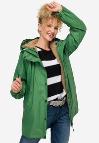 Giacca impermeabile verde con cappuccio, dotata di fodera in peluche beige, bottoni a pressione e polsini elasticizzati, indossata sopra una maglietta a righe bianco e nero.