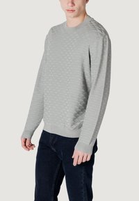 Lichtgrijze gebreide trui met een gestructureerd ruitpatroon, geribde manchetten en zoom, gecombineerd met donkere jeans.