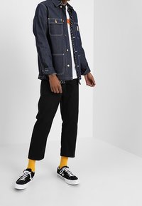 Jeansjacka med två framfickor och knappstängning, lagd över en vit t-shirt med svart prickmönster. Svarta byxor och svarta sneakers med vita detaljer.