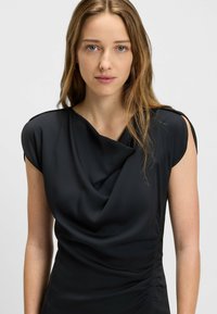 Schwarzes ärmelloses Kleid mit drapiertem Ausschnitt, glatter Textur und Raffung seitlich; weicher Stoff mit leichtem Schimmer.