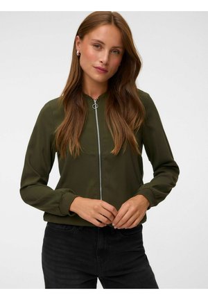 Femme aux longs cheveux bruns portant une veste zippée vert olive et un pantalon noir, debout sur un fond blanc uni.