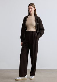 Veste bomber marron côtelée avec passepoil blanc, haut côtelé beige ajusté, pantalon taille haute foncé et baskets argentées avec semelles beiges.