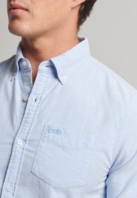 Lichtblauw overhemd met knopen, gemaakt van zachte stof, met een zak en een klein blauw merkje op de borst. Klassiek kraagontwerp.