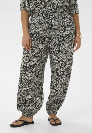 Luźne czarne spodnie z misternymi białymi kwiatowymi i paisley wzorami, elastyczne u dołu nogawki, noszone z czarnymi klapkami i złotą bransoletką na nadgarstku.