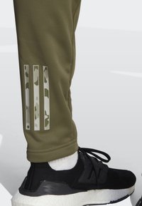 Grønne træningsbukser med tekstureret stof har tre vertikale hvide og camouflagemønstrede striber. Sorte sneakers med hvid sål og synlige sokker.