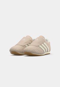 Beige krāsas Adidas sporta apavi ar baltām svītrām un gumijas zole, ar platu velcro siksnu pāri augšai.