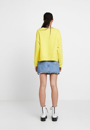 Mujer con cabello trenzado, vistiendo un sudadera amarilla, falda de mezclilla azul y botas blancas hasta el tobillo, de pie de espaldas sobre un fondo liso.