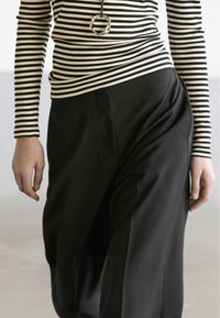 Schwarze Hose mit lockerem Schnitt, kombiniert mit einem schwarz-weiß gestreiften Langarmshirt. Auffälliges goldenes, rundes Halsketten-Accessoire.