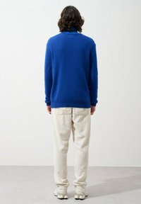 Blauer Wollpullover mit hohem Kragen, gerippten Bündchen, kombiniert mit creamfarbenen Cordhosen und grauen Sneakern, von hinten gesehen.