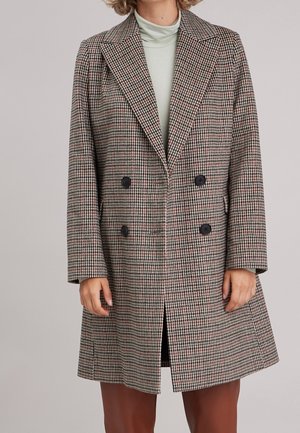 Femme portant un manteau croisé en pied-de-poule avec des boutons noirs sur un col roulé vert clair et un pantalon marron.