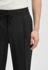 Black trousers with an elastic waistband and drawstring, featuring a smooth texture and front creases. Hand positioned at the side.