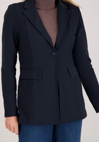 Marineblazer met een getailleerde pasvorm, één knoopsluiting, ingezette revers en twee steekzakken aan de voorkant, gecombineerd met een bruine coltrui eronder.